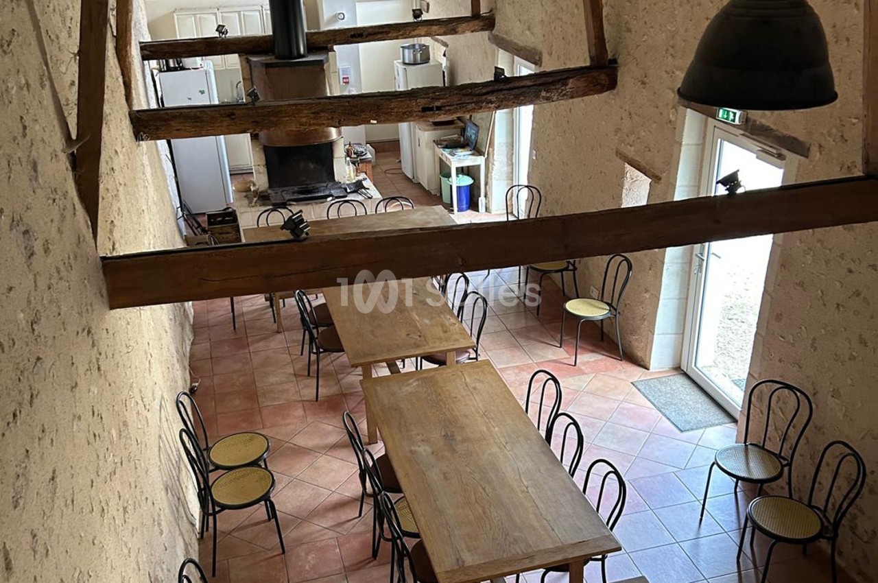Salle à manger rustique avec tables en bois, chaises en métal et poutres apparentes, vue depuis une mezzanine.