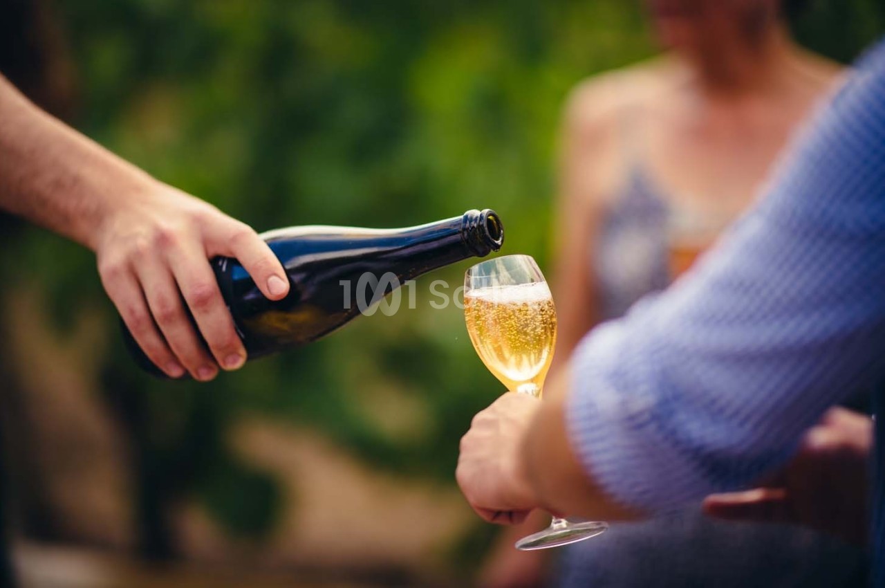 Une main verse du vin pétillant dans un verre tenu par une autre personne, en extérieur avec un arrière-plan flou.