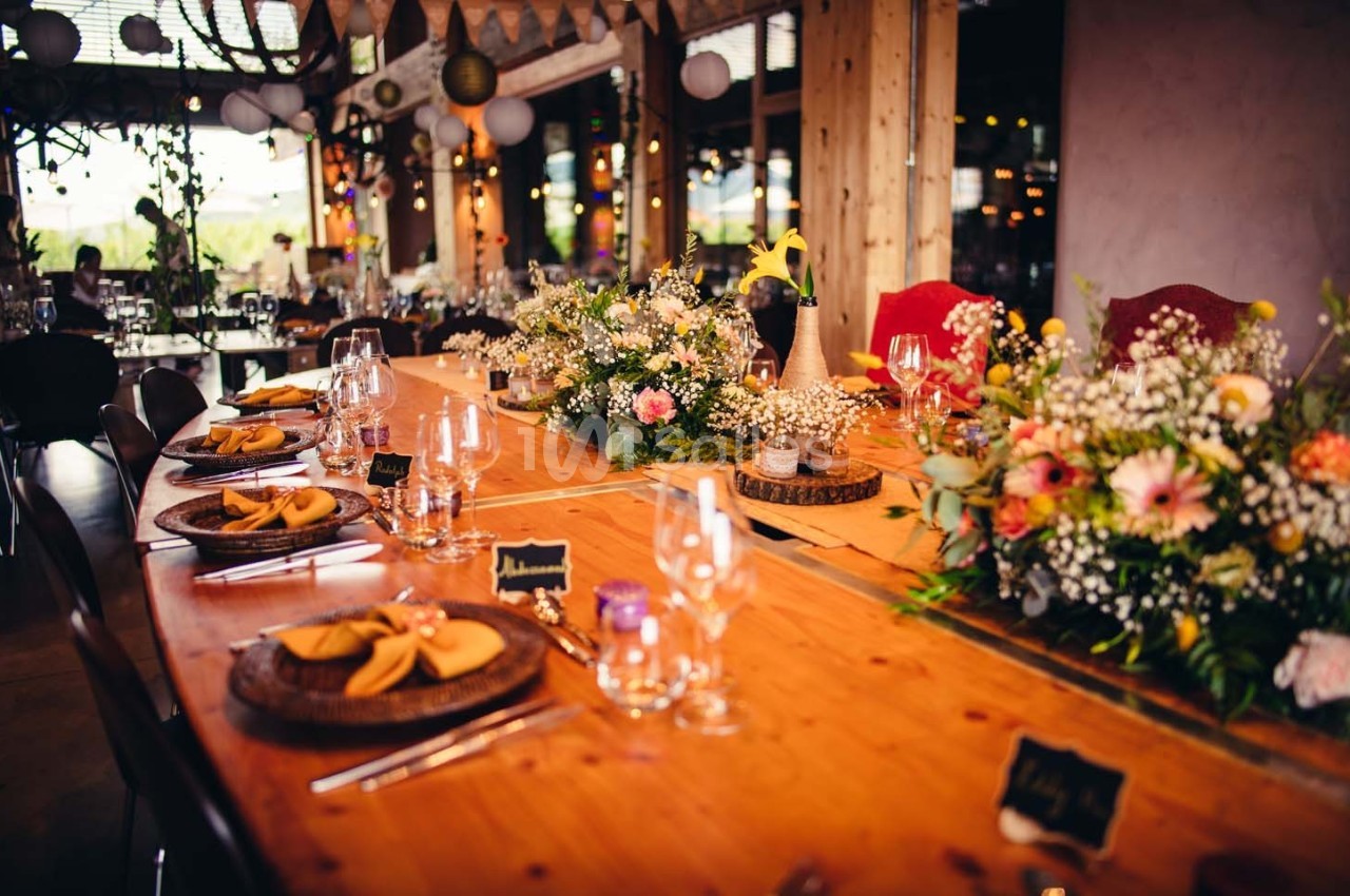 Table en bois décorée pour un événement, avec fleurs, assiettes dressées et verres dans une ambiance festive.