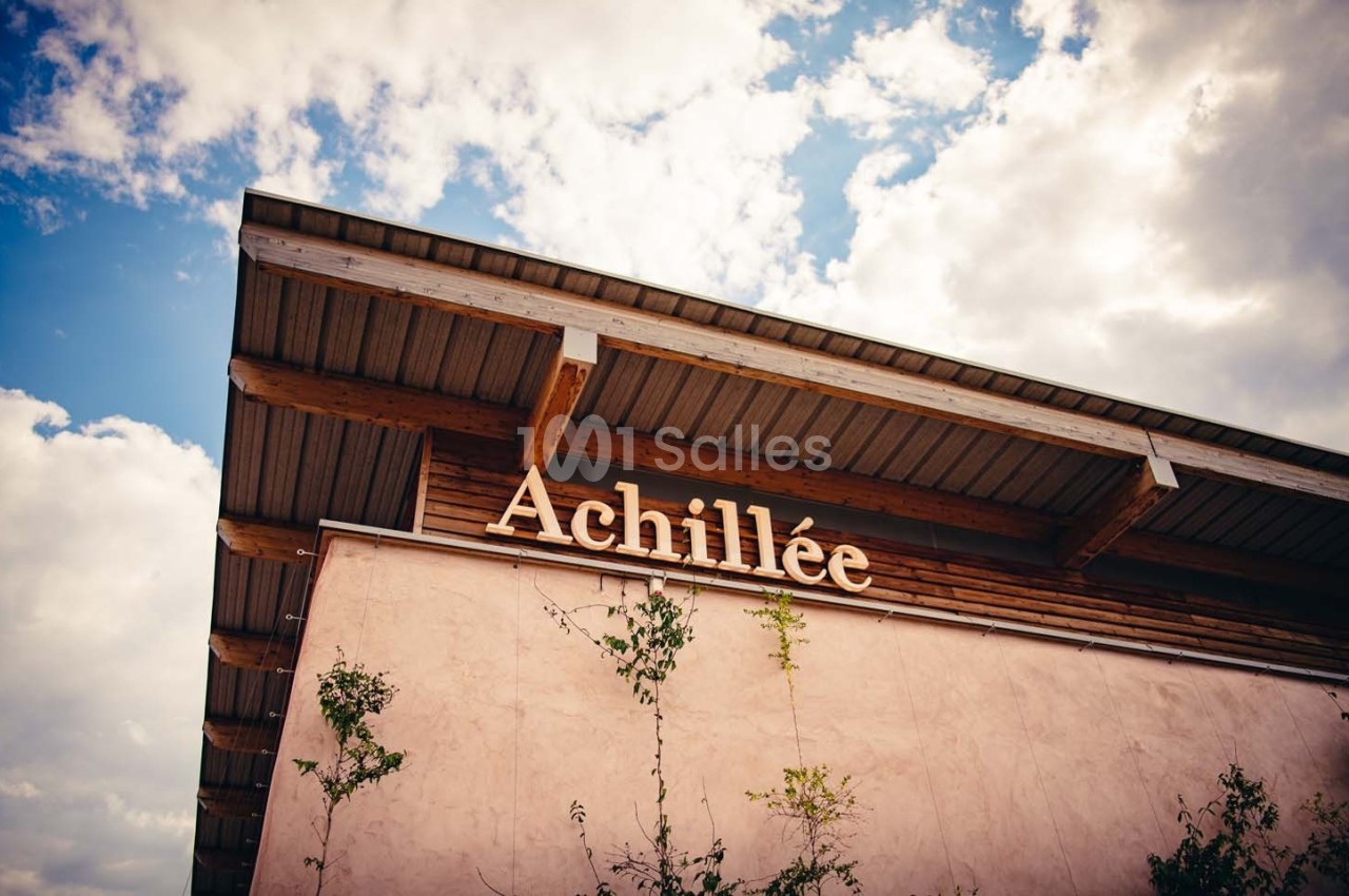 Façade d'un bâtiment en bois et crépi rose avec le mot ’Achillée’, sous un ciel partiellement nuageux.