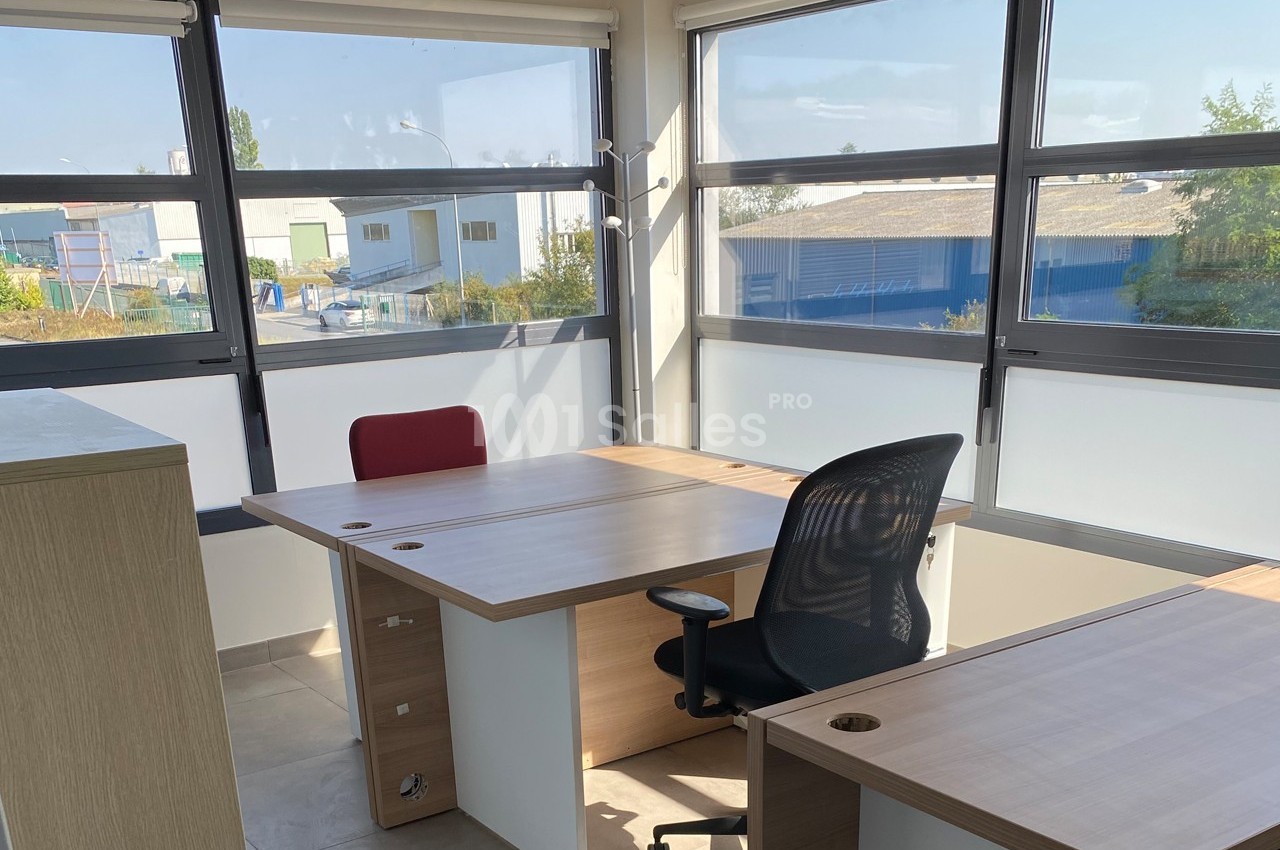 Bureau lumineux avec deux postes de travail, chaises et grandes fenêtres offrant une vue sur des bâtiments industriels.