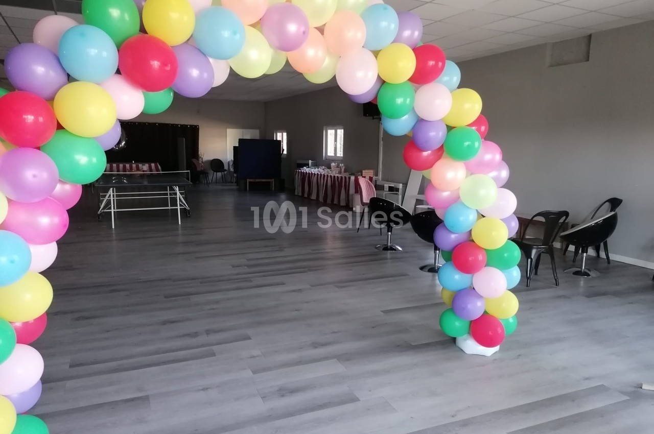 Arche de ballons multicolores dans une salle vide avec des tables et des chaises en arrière-plan.
