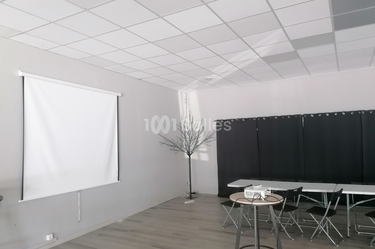 Salle lumineuse avec sol en bois, écran de projection, tables, chaises pliantes et rideaux noirs.