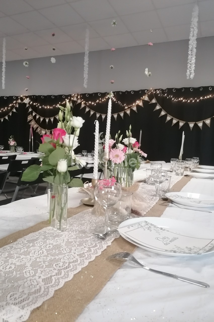 Salle décorée pour un événement avec tables dressées, nappes blanches, fleurs, bougies et guirlandes lumineuses.