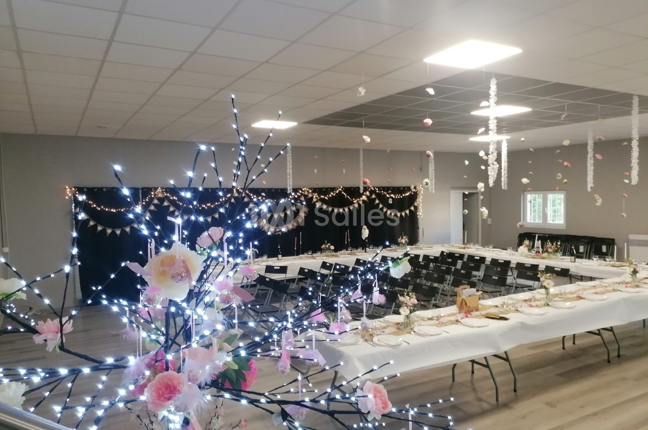 Salle décorée pour un événement avec tables dressées, guirlandes lumineuses, fleurs et ornements suspendus.