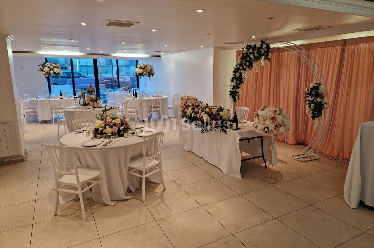 Salle de réception décorée avec des tables rondes, nappes blanches, compositions florales et arche ornée de fleurs.