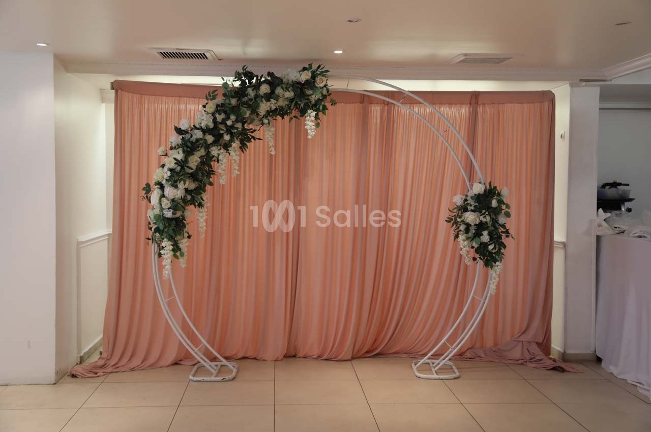 Arche blanche décorée de fleurs blanches et vertes devant un rideau rose dans une salle lumineuse.