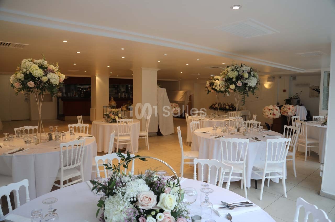 Salle de réception lumineuse avec tables rondes décorées de fleurs blanches et roses, chaises blanches et éclairage doux.
