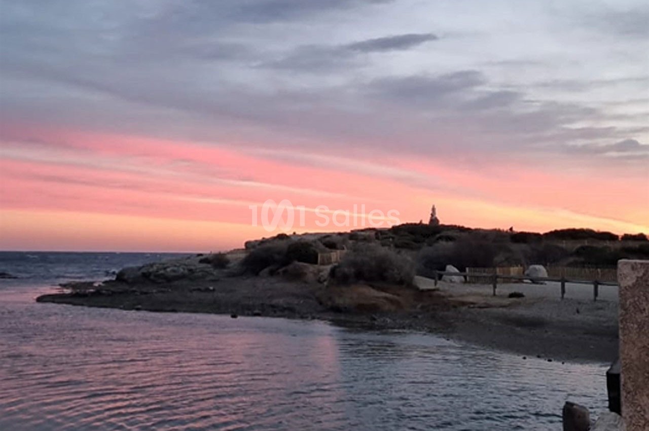 Coucher de soleil aux teintes roses et violettes sur une plage avec une petite colline et un bâtiment en arrière-plan.