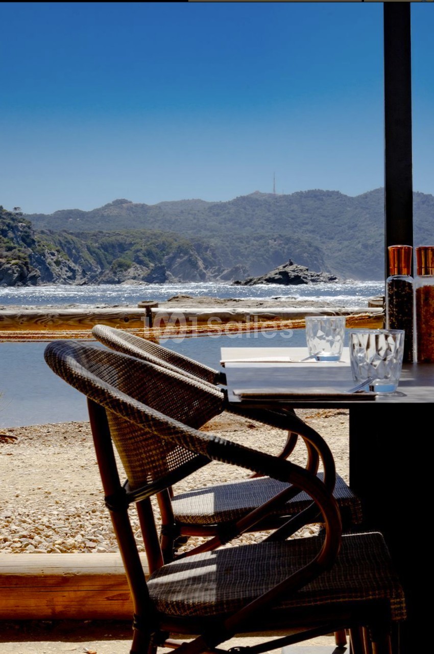 Chaises en osier et table dressée avec vue sur une plage, des collines et l'océan sous un ciel dégagé.