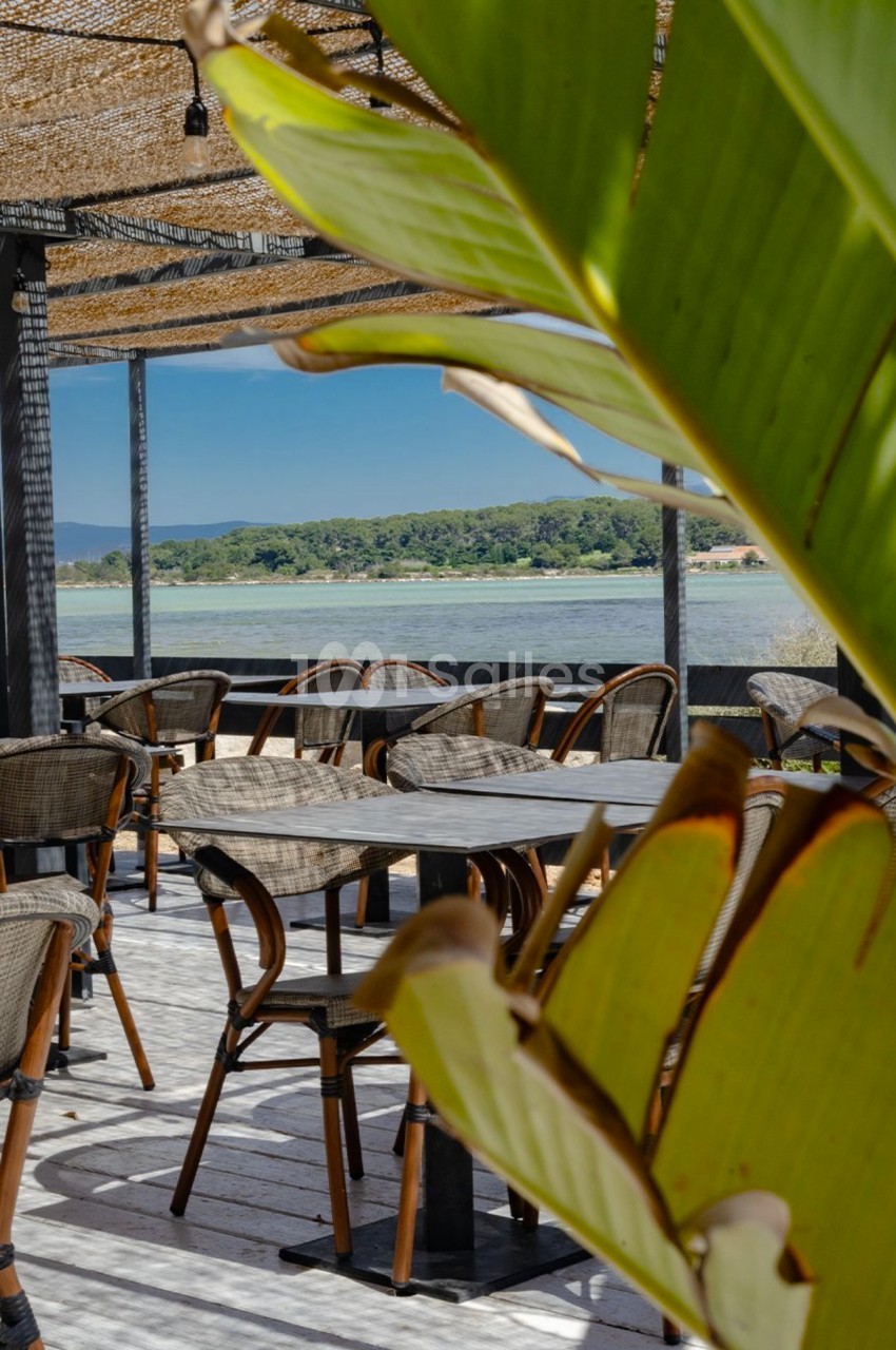 Terrasse extérieure avec des tables et chaises en rotin, vue sur un paysage de lac et végétation tropicale au premier plan.