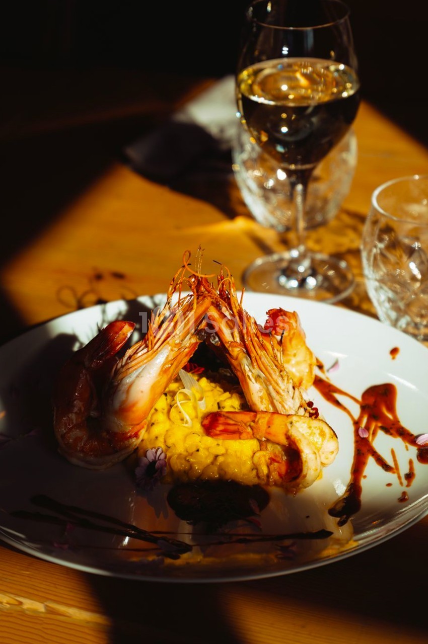 Assiette de risotto au safran avec crevettes grillées, décorée de sauce, servie sur une table en bois avec un verre de vin.