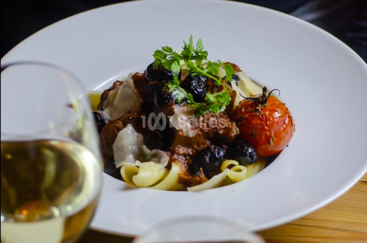 Assiette de pâtes garnie de viande en sauce, olives noires, tomate rôtie et herbes, servie avec du vin blanc.