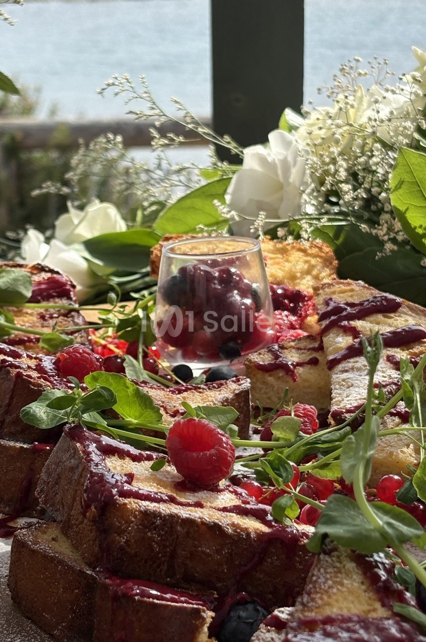 Tranches de brioche dorées garnies de fruits rouges, nappées de coulis, entourées de verdure et de fleurs, en extérieur.