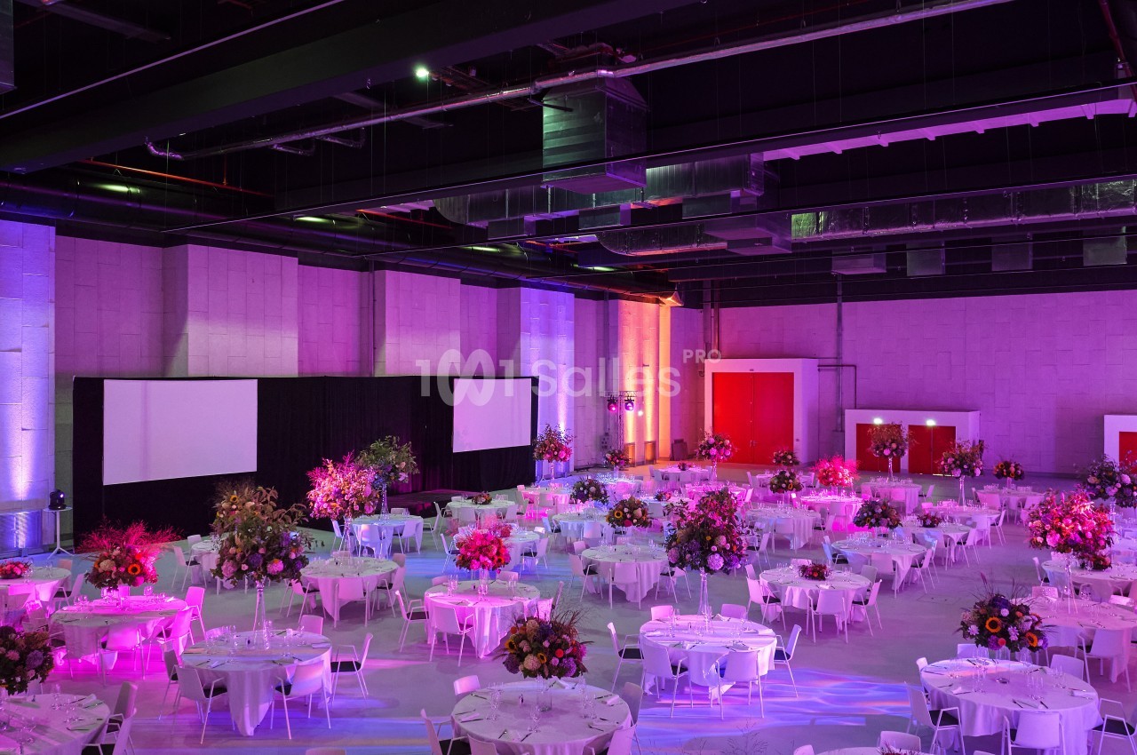 Salle de réception décorée avec des tables rondes, nappes blanches et arrangements floraux sous un éclairage coloré.