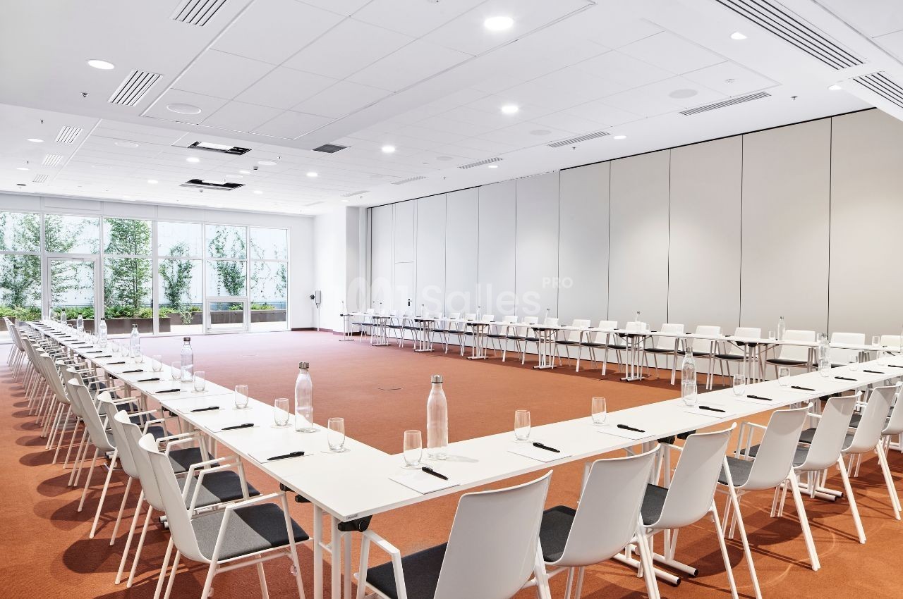 Salle de conférence lumineuse avec tables disposées en U, chaises blanches et grandes baies vitrées donnant sur un jardin.