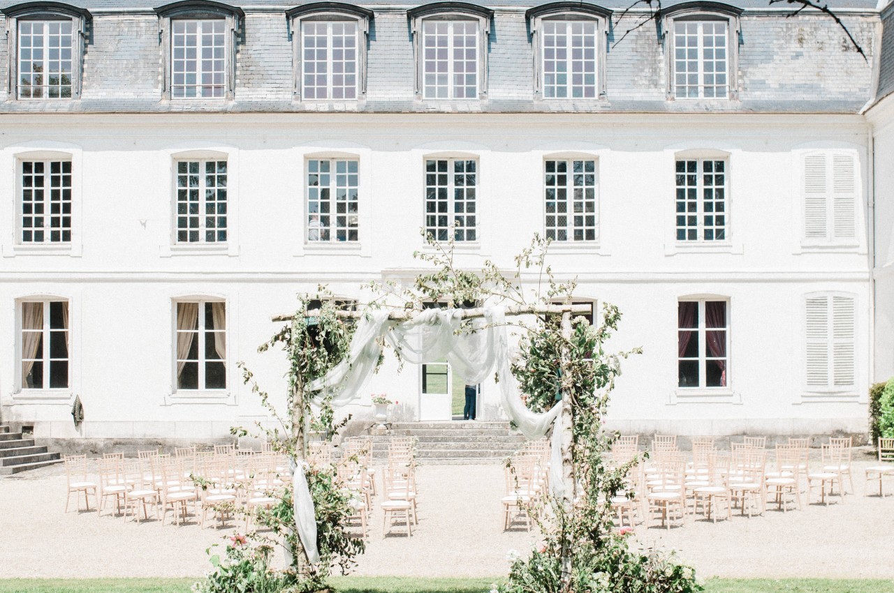Façade d'un bâtiment blanc de style classique avec une arche décorée et des chaises disposées en extérieur.