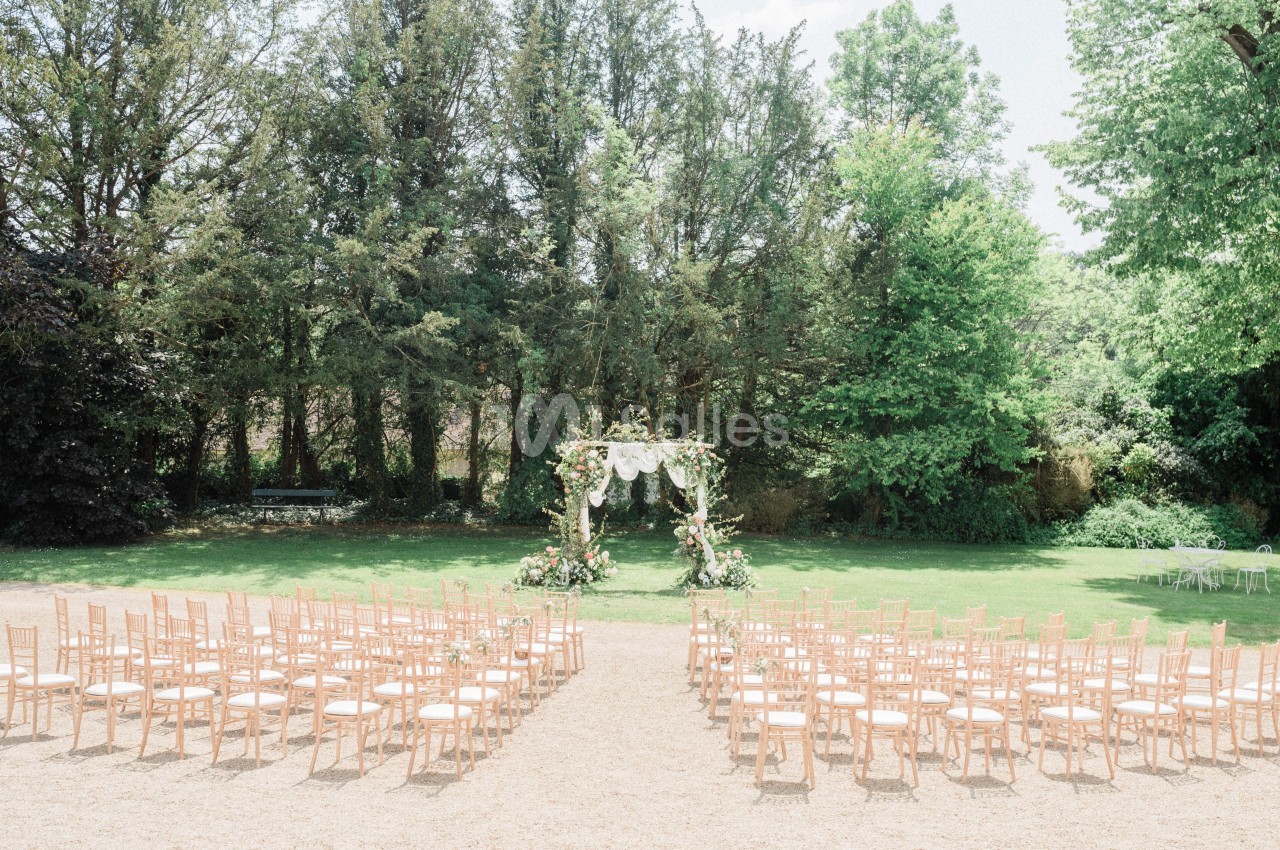 Chaises disposées en rangées face à une arche décorée de fleurs dans un jardin verdoyant pour une cérémonie en plein air.