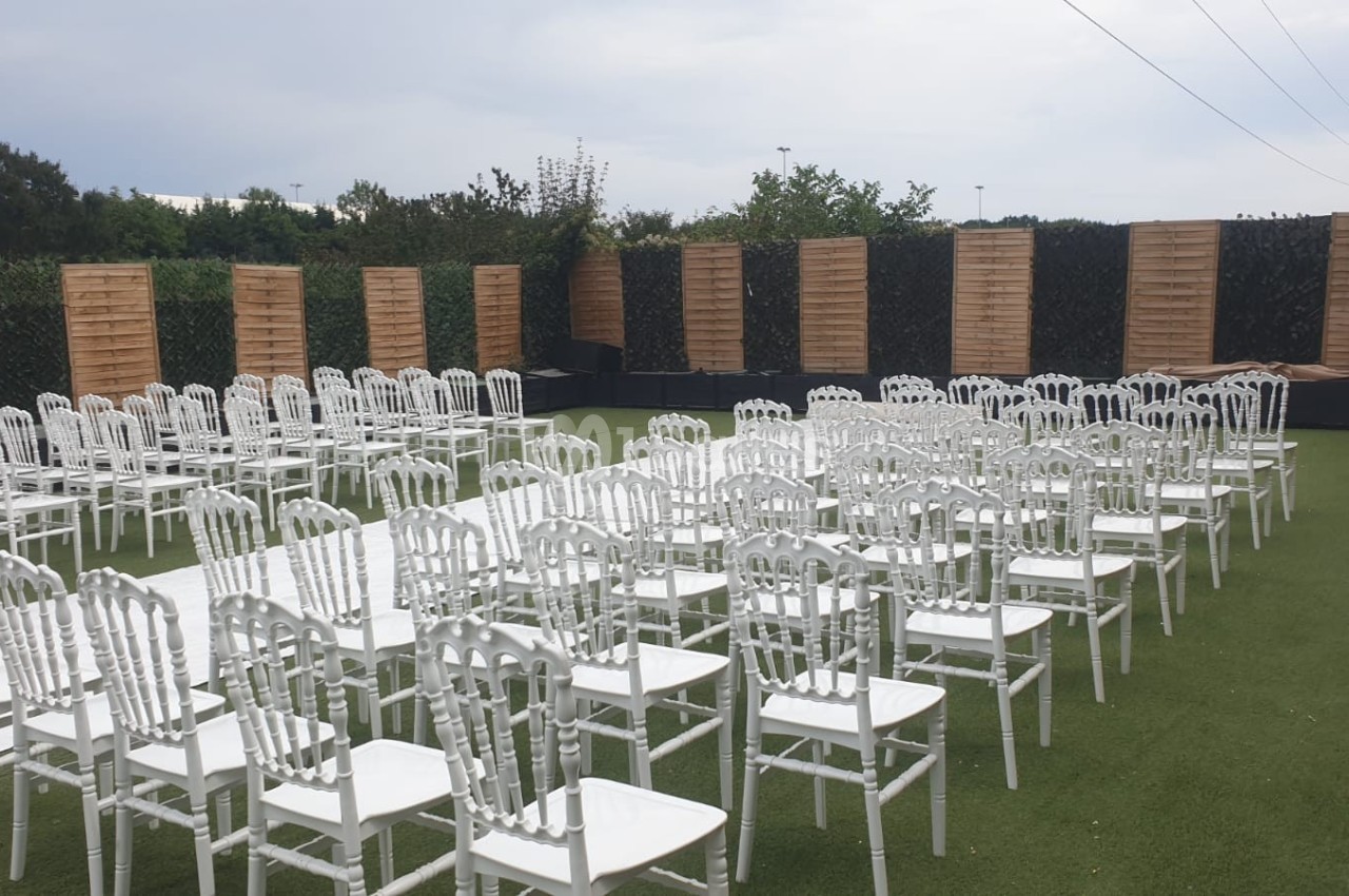 Chaises blanches alignées en rangées sur une pelouse artificielle, entourées de panneaux en bois et de verdure.