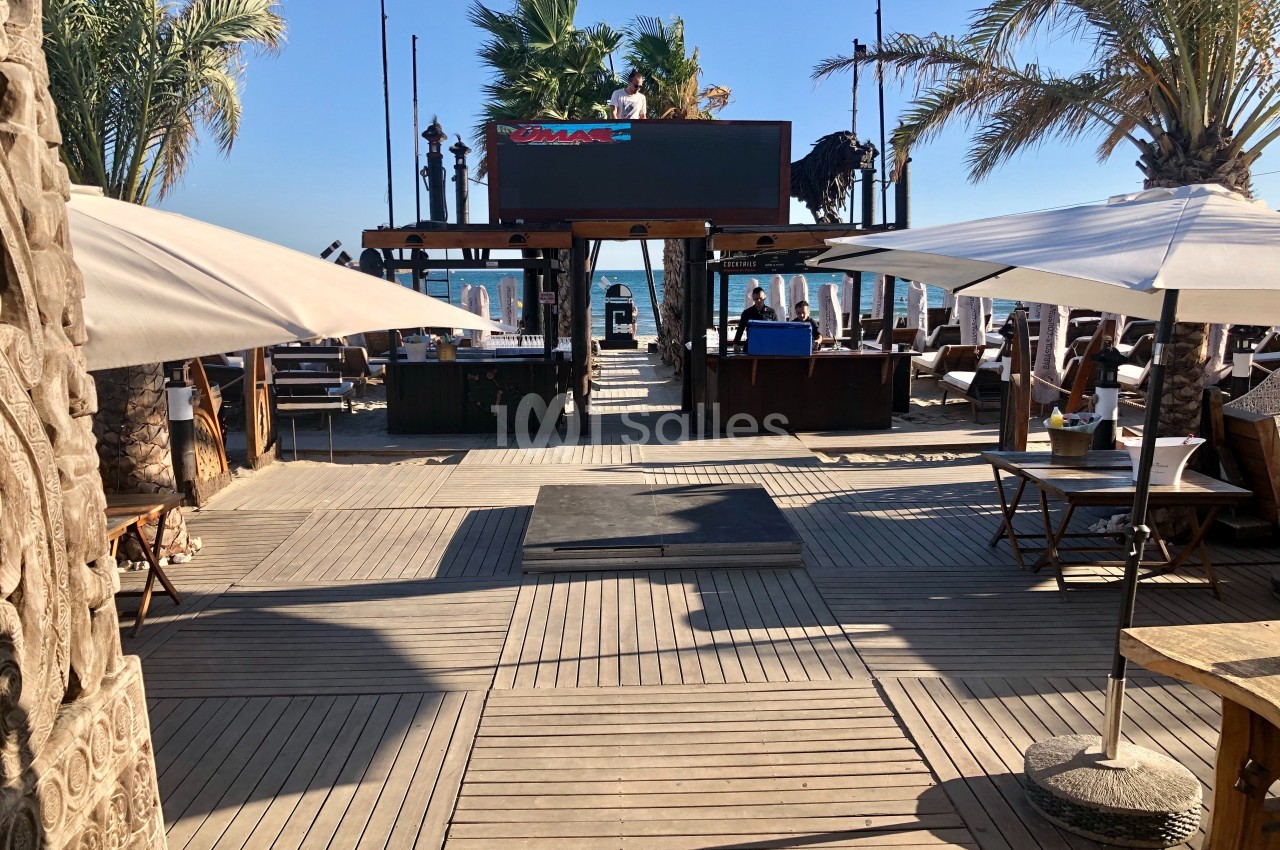 Terrasse en bois avec transats, parasols et palmiers, donnant sur la mer sous un ciel bleu clair.