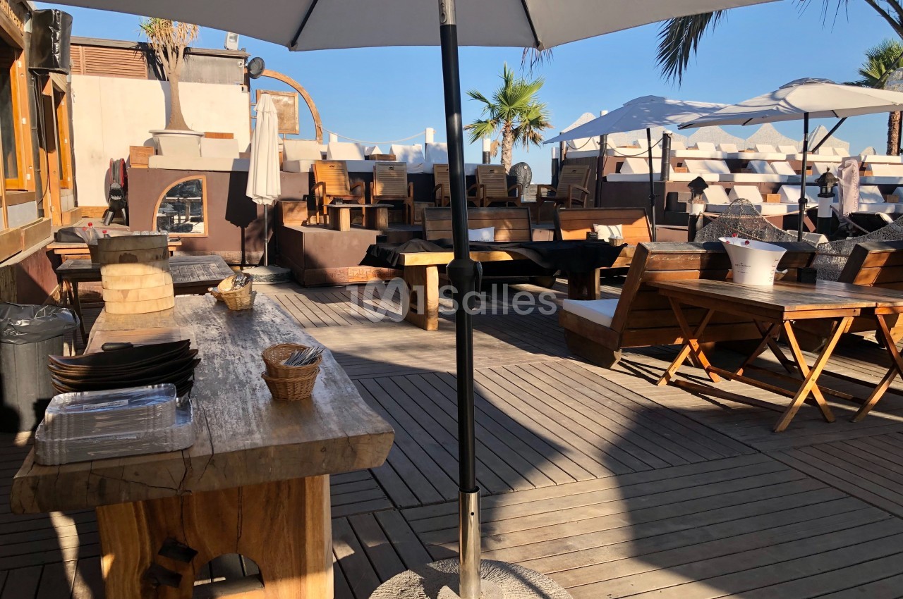 Terrasse en bois avec tables et chaises, parasols ouverts, et vue sur des palmiers sous un ciel dégagé.