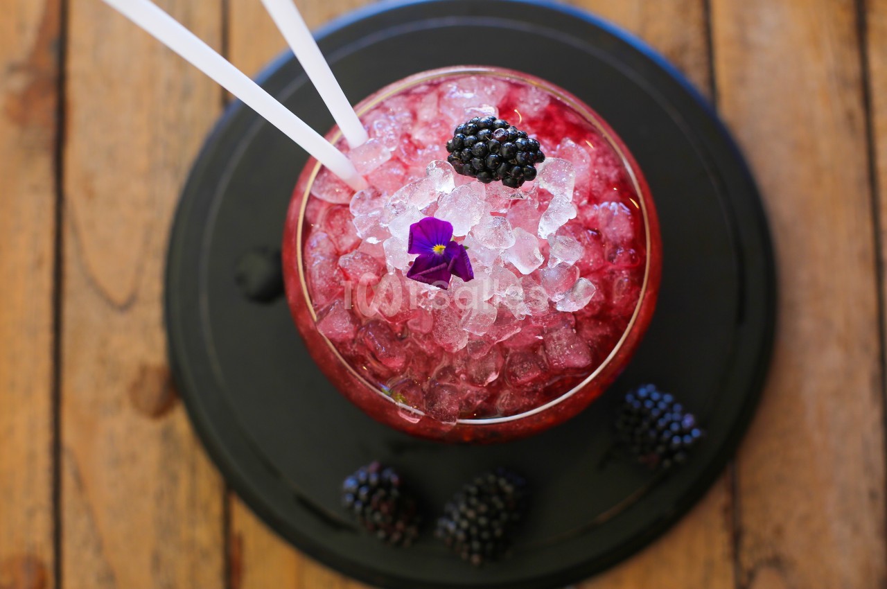 Verre de cocktail rouge avec glace pilée, mûres fraîches et une fleur violette, posé sur une table en bois.