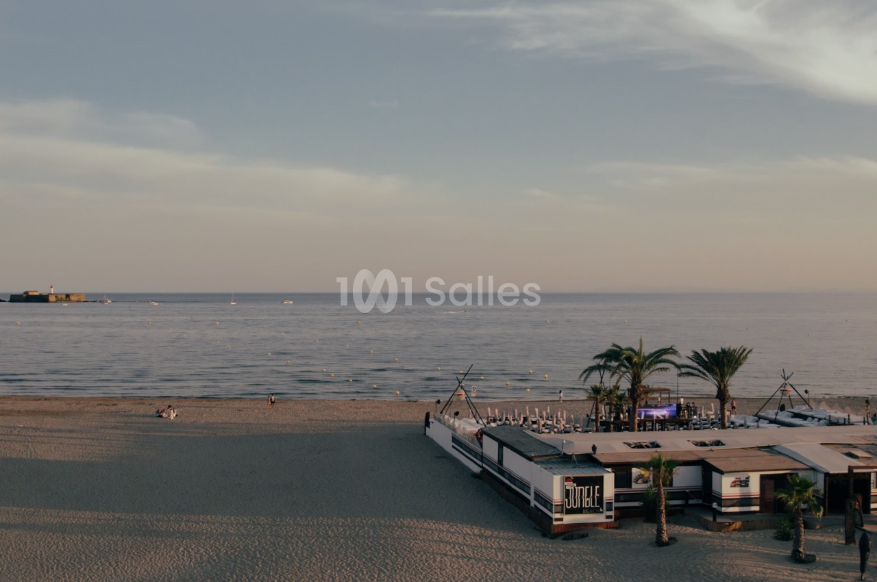 Plage au coucher du soleil avec un restaurant en bord de mer, quelques palmiers et un fort visible au loin sur l'eau.