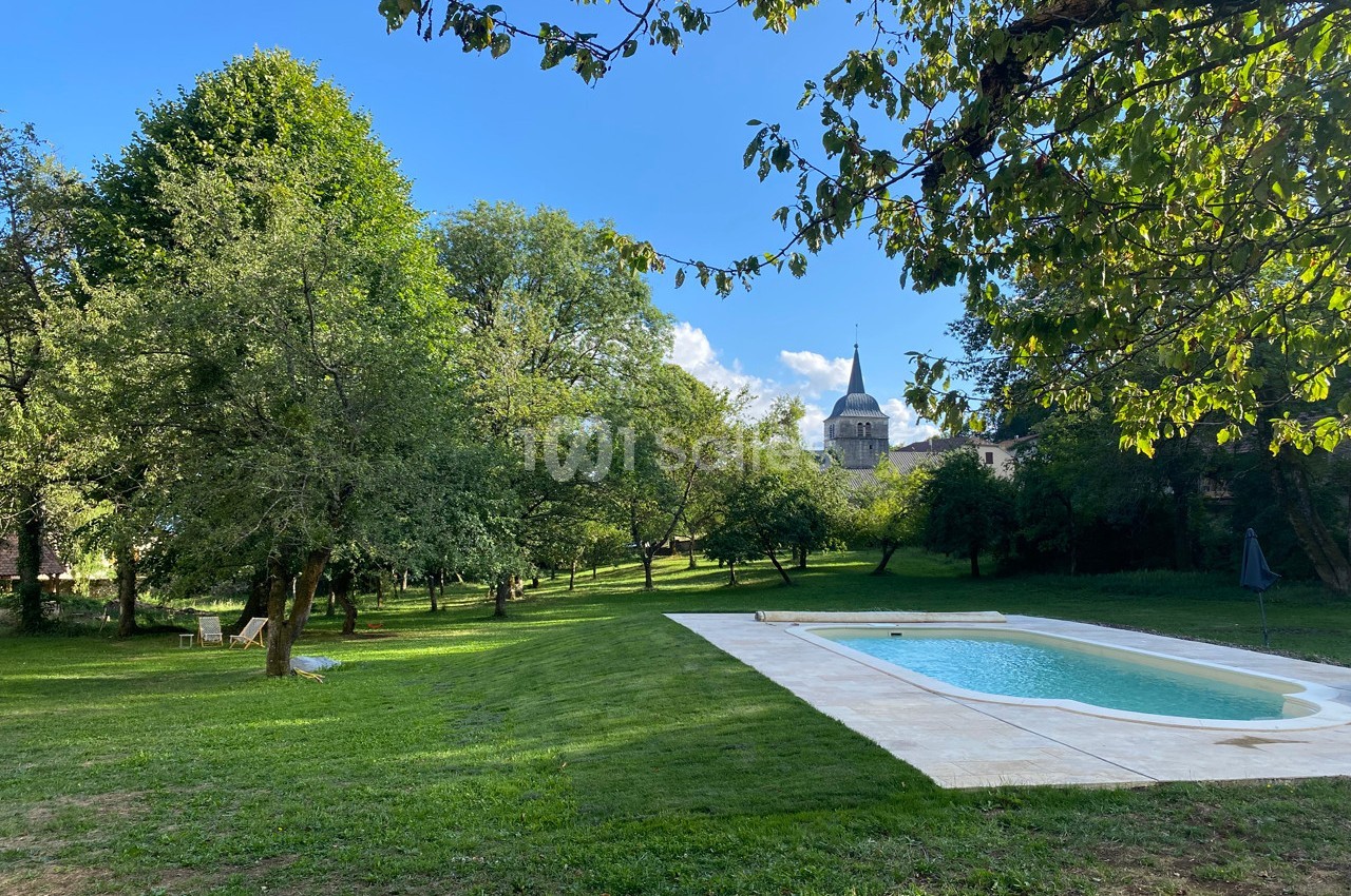 Jardin verdoyant avec piscine rectangulaire, arbres ombragés et clocher d'église visible en arrière-plan.