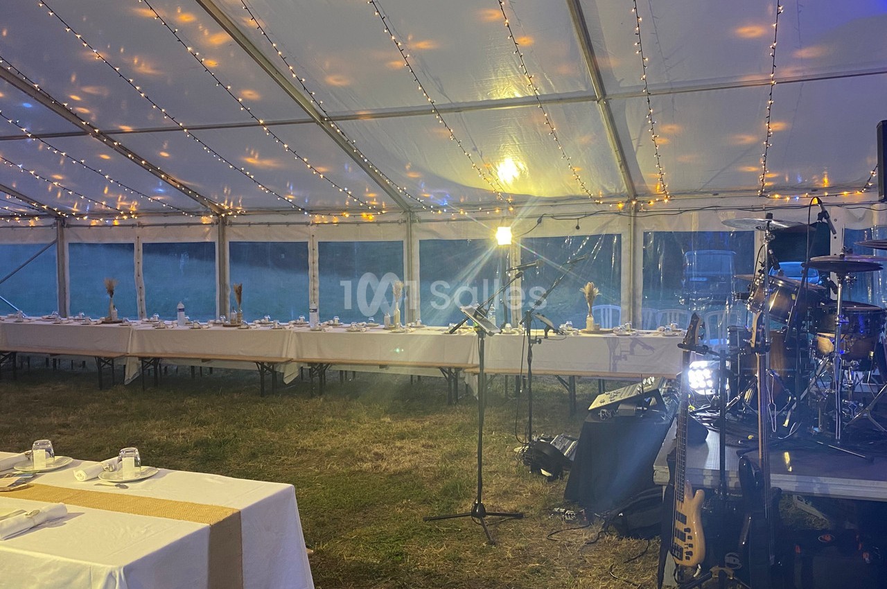 Intérieur d'une tente éclairée avec des guirlandes lumineuses, tables dressées et scène équipée d'instruments de musique.