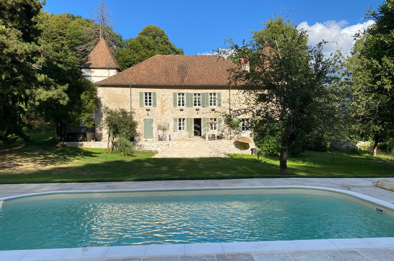 Façade d'une maison en pierre avec toit en tuiles, entourée d'arbres, vue depuis une piscine.