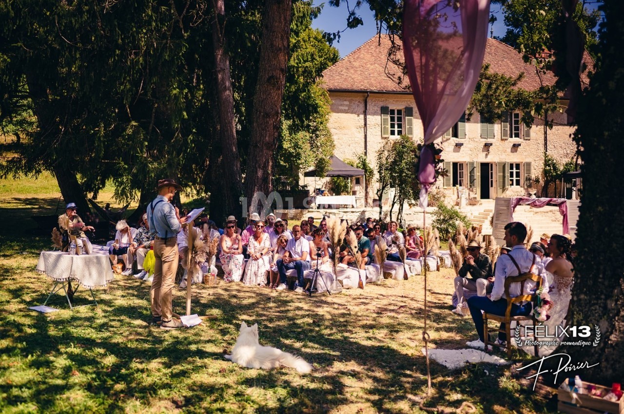 Cérémonie en plein air avec des invités assis sous des arbres, devant une maison en pierre.