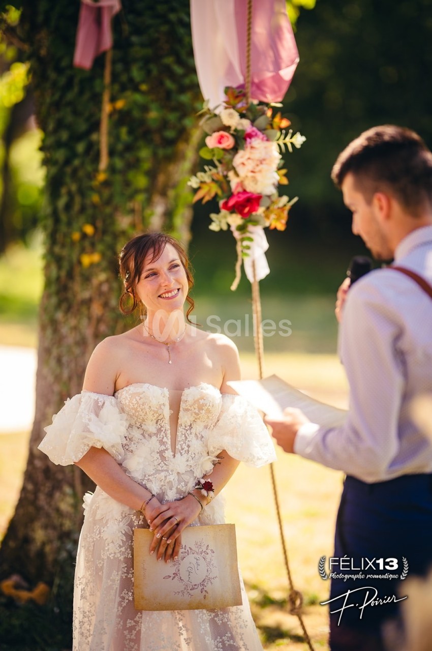 Une mariée souriante écoute un discours du marié lors d'une cérémonie en extérieur, près d'un arbre décoré.