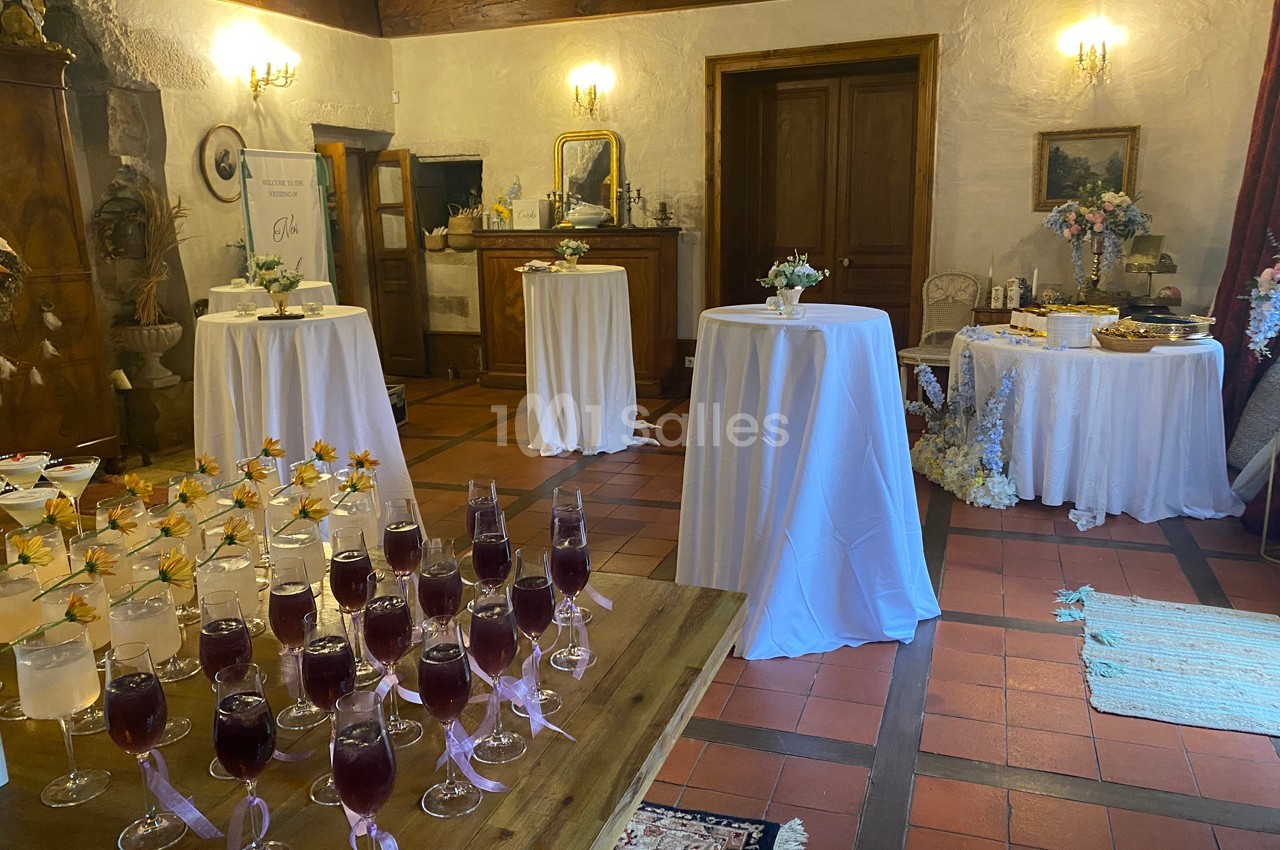 Salle décorée pour un événement avec tables hautes, nappes blanches, fleurs et verres de cocktail sur une table en bois.