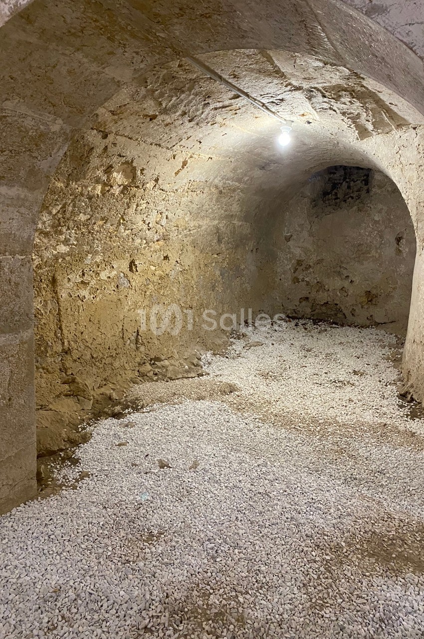 Cave voûtée en pierre avec sol recouvert de gravier, éclairée par une ampoule suspendue.