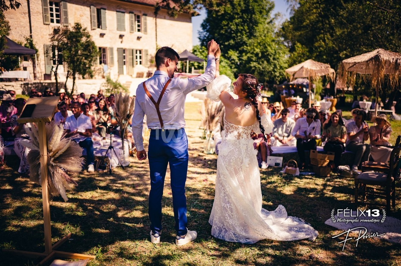 Un couple danse en plein air lors d'une cérémonie de mariage, entouré d'invités assis devant une maison en pierre.
