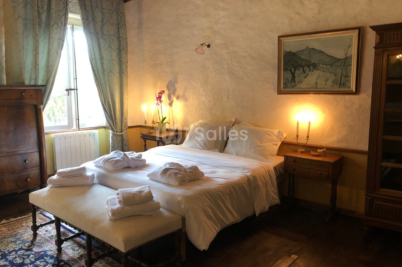 Chambre lumineuse avec lit double, draps blancs, bougies allumées, orchidée sur une table et fenêtre donnant sur l'extérieur.