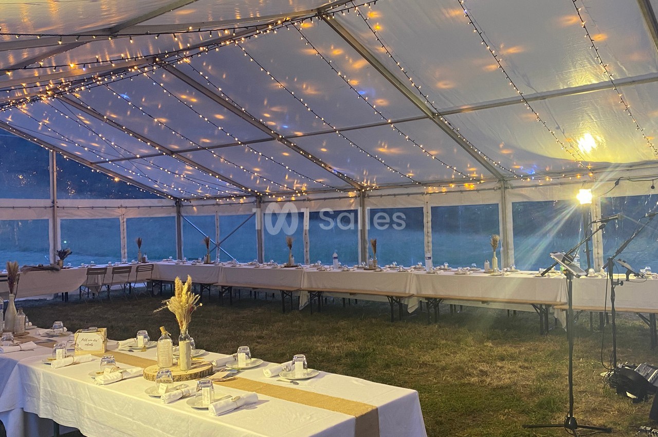 Grande tente éclairée par des guirlandes lumineuses, aménagée avec des tables décorées pour un événement en soirée.