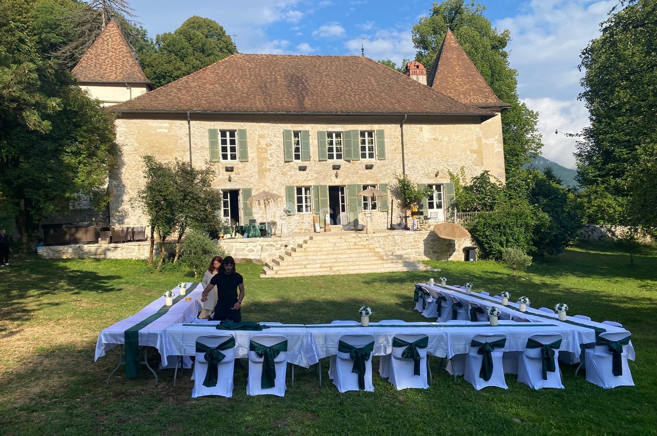 Tables dressées en extérieur devant une grande maison en pierre entourée de verdure, par une journée ensoleillée.