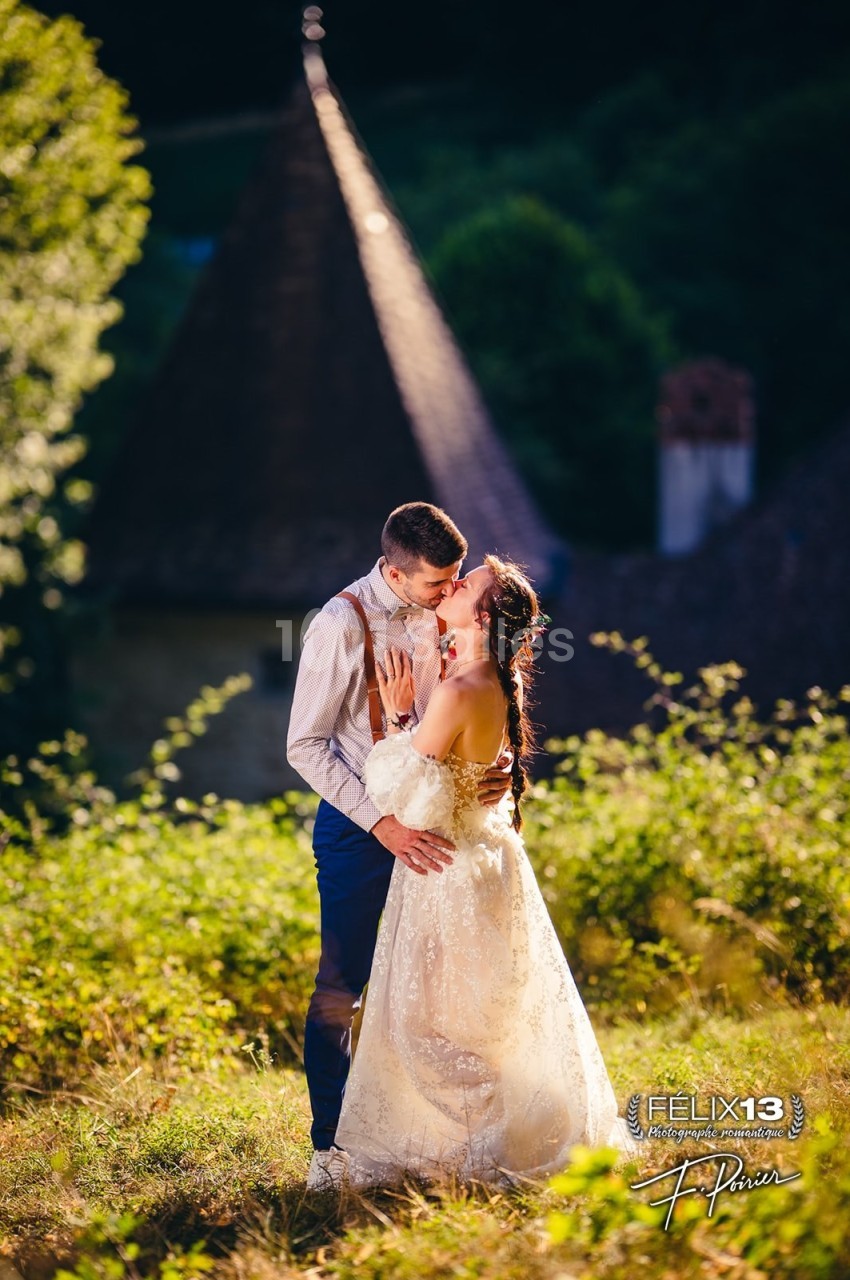 Un couple en tenue de mariage s'embrasse dans un jardin ensoleillé devant un bâtiment au toit en pointe.