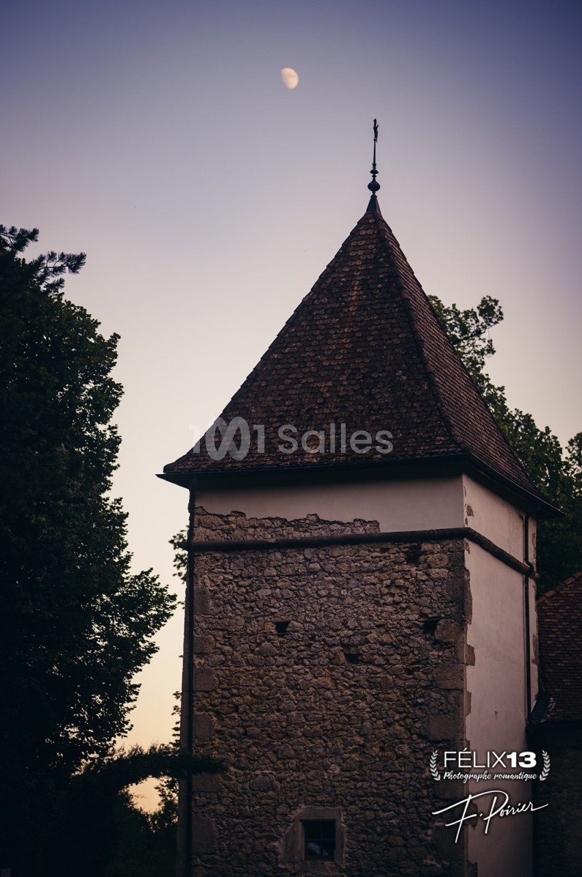 Tour en pierre avec un toit en tuiles sous un ciel crépusculaire, entourée d'arbres et une lune visible en arrière-plan.