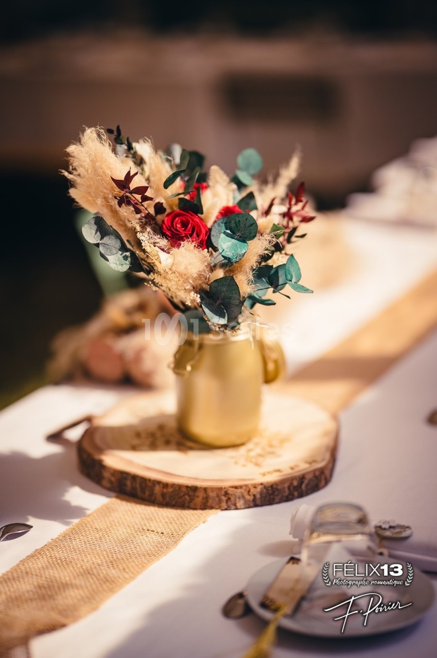 Bouquet de fleurs séchées et rouges dans un vase doré, posé sur une rondelle de bois, décorant une table élégante.