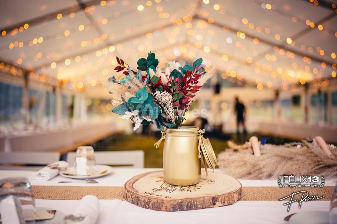 Bouquet de fleurs séchées dans un vase doré sur une table en bois, sous une tente éclairée de guirlandes lumineuses.