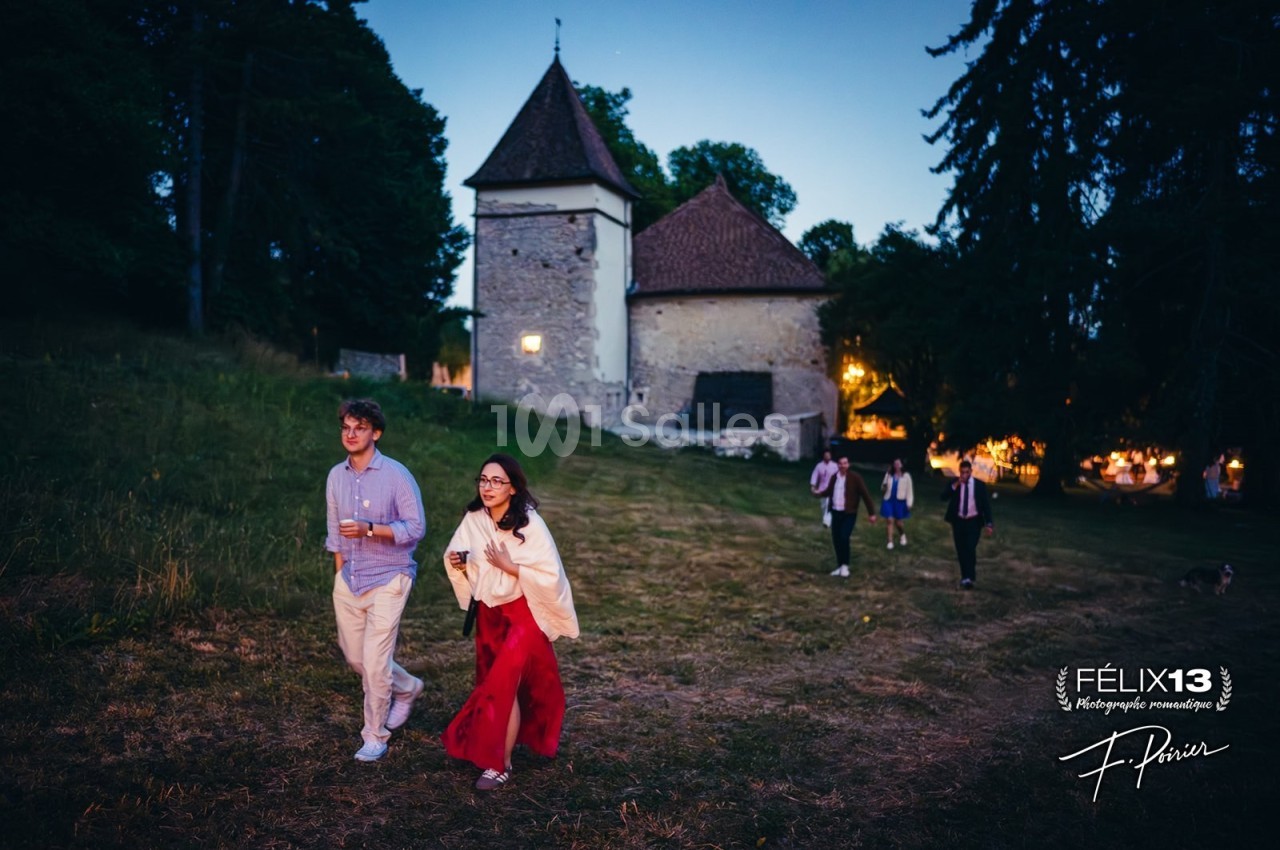 Un couple marche sur une pelouse au crépuscule devant un bâtiment en pierre éclairé, suivi par d'autres personnes.