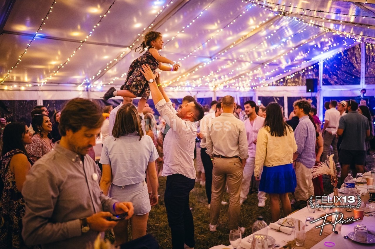 Un homme soulève une enfant dans ses bras au milieu d'une fête sous une tente illuminée de guirlandes.