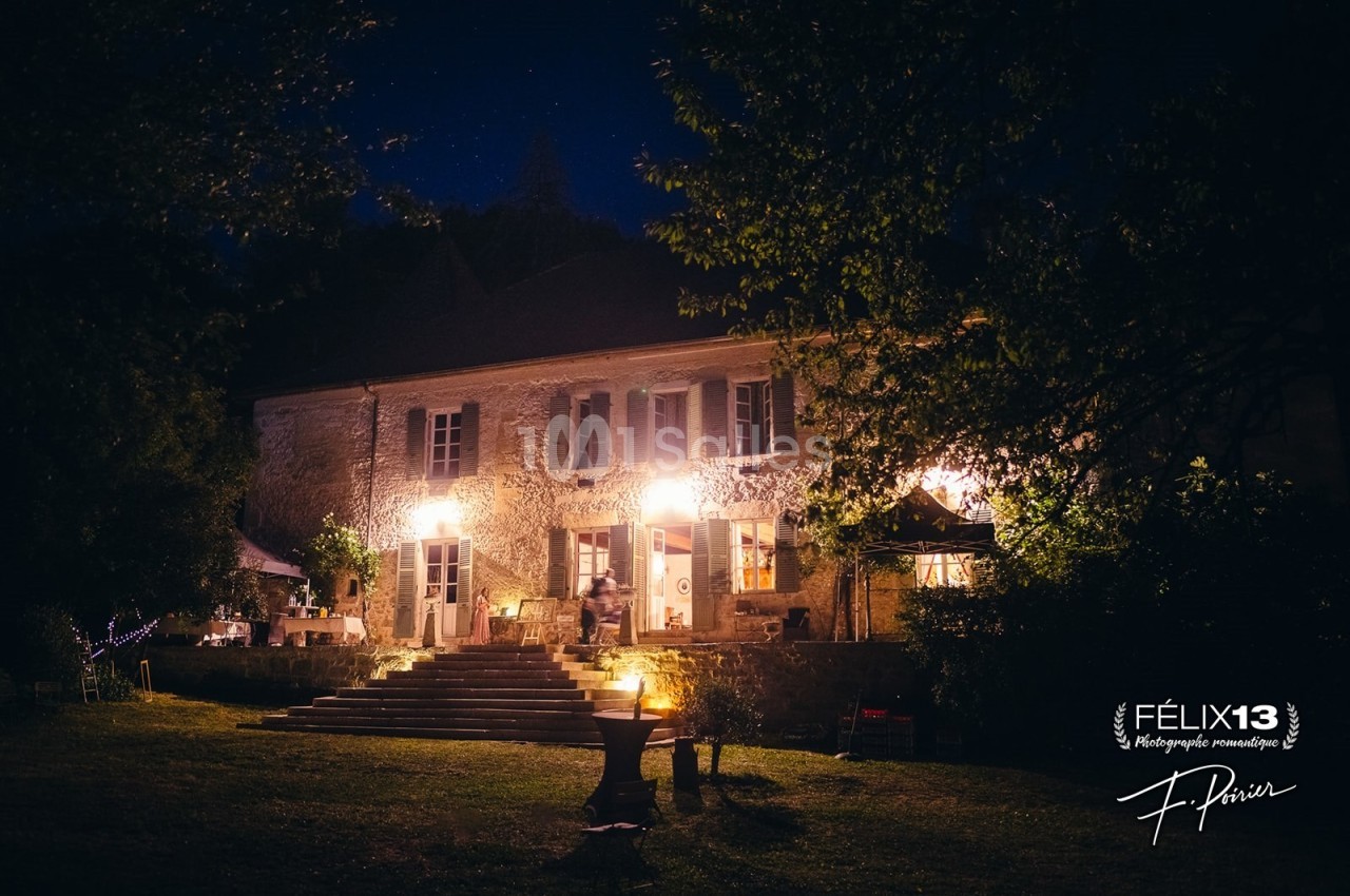 Façade d'une maison en pierre éclairée la nuit, entourée d'arbres et d'un jardin avec des lumières tamisées.