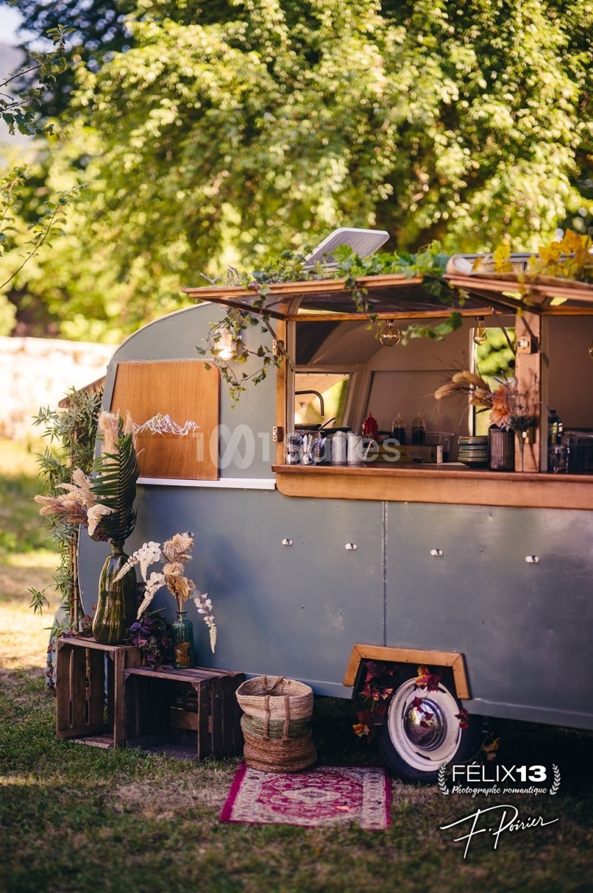 Caravane vintage aménagée en bar, entourée de décorations florales et de caisses en bois, dans un cadre extérieur verdoyant.