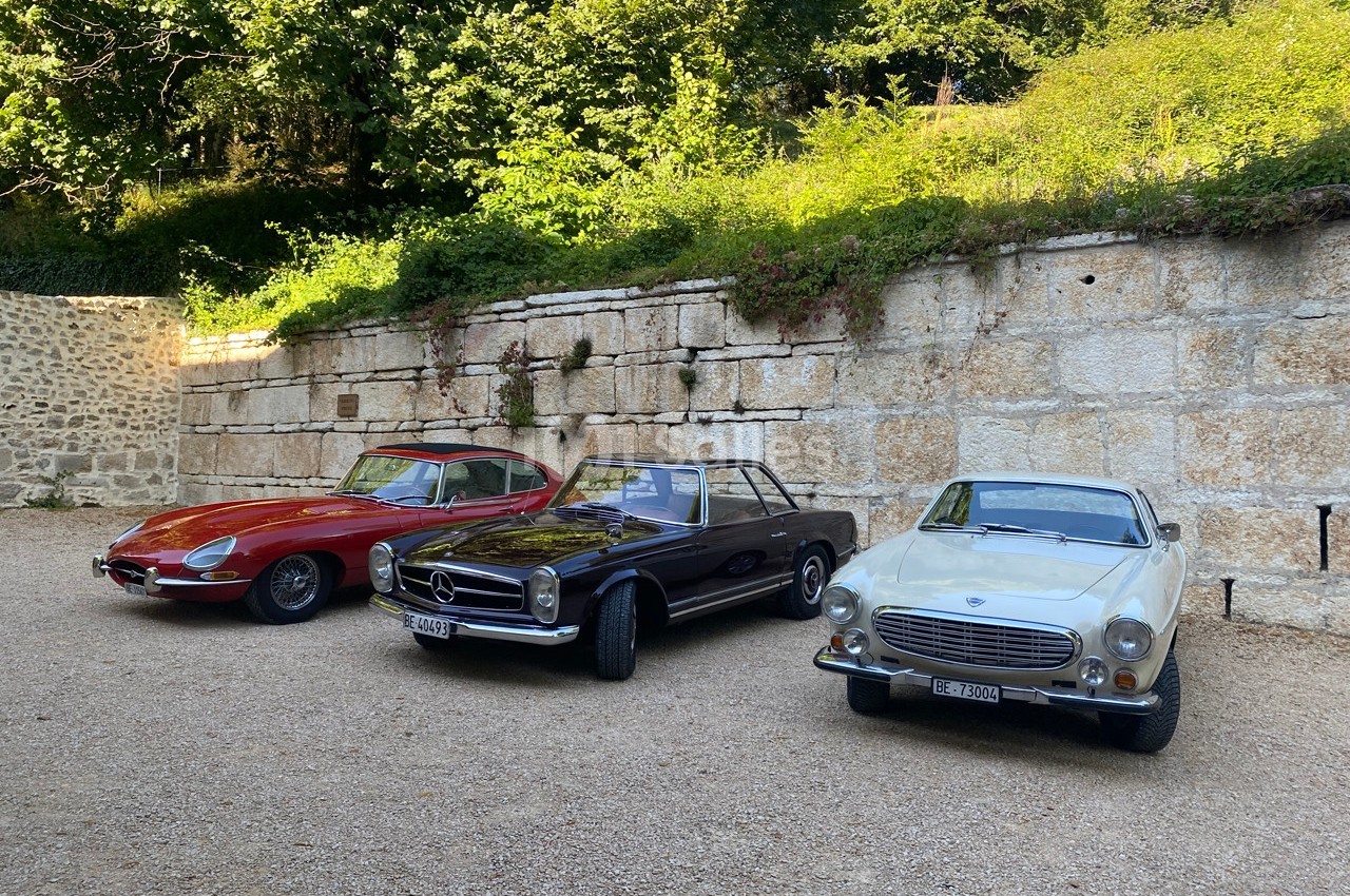 Trois voitures anciennes stationnées devant un mur de pierre, entourées de végétation.