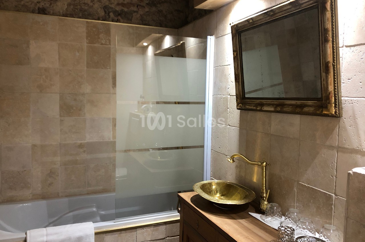 Salle de bain avec baignoire, paroi vitrée, vasque dorée sur meuble en bois et miroir encadré au mur.
