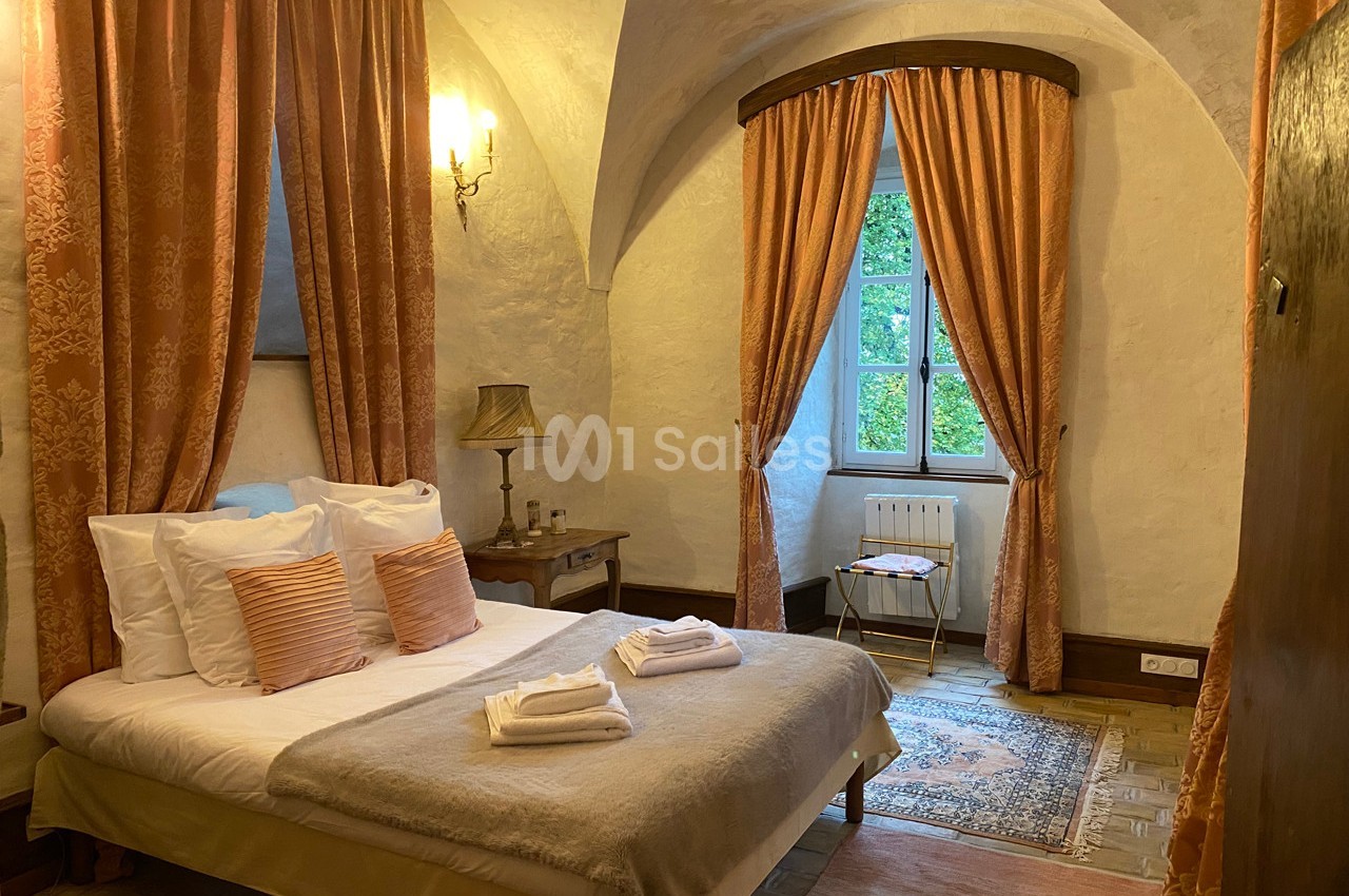 Chambre avec lit double, rideaux orange, murs en pierre, lampe de chevet et fenêtre donnant sur un jardin verdoyant.
