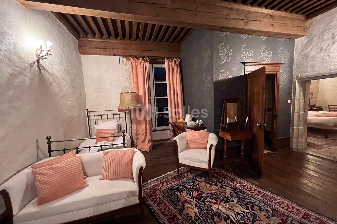 Salon rustique avec poutres apparentes, mobilier ancien, tapis coloré, rideaux roses et éclairage doux.